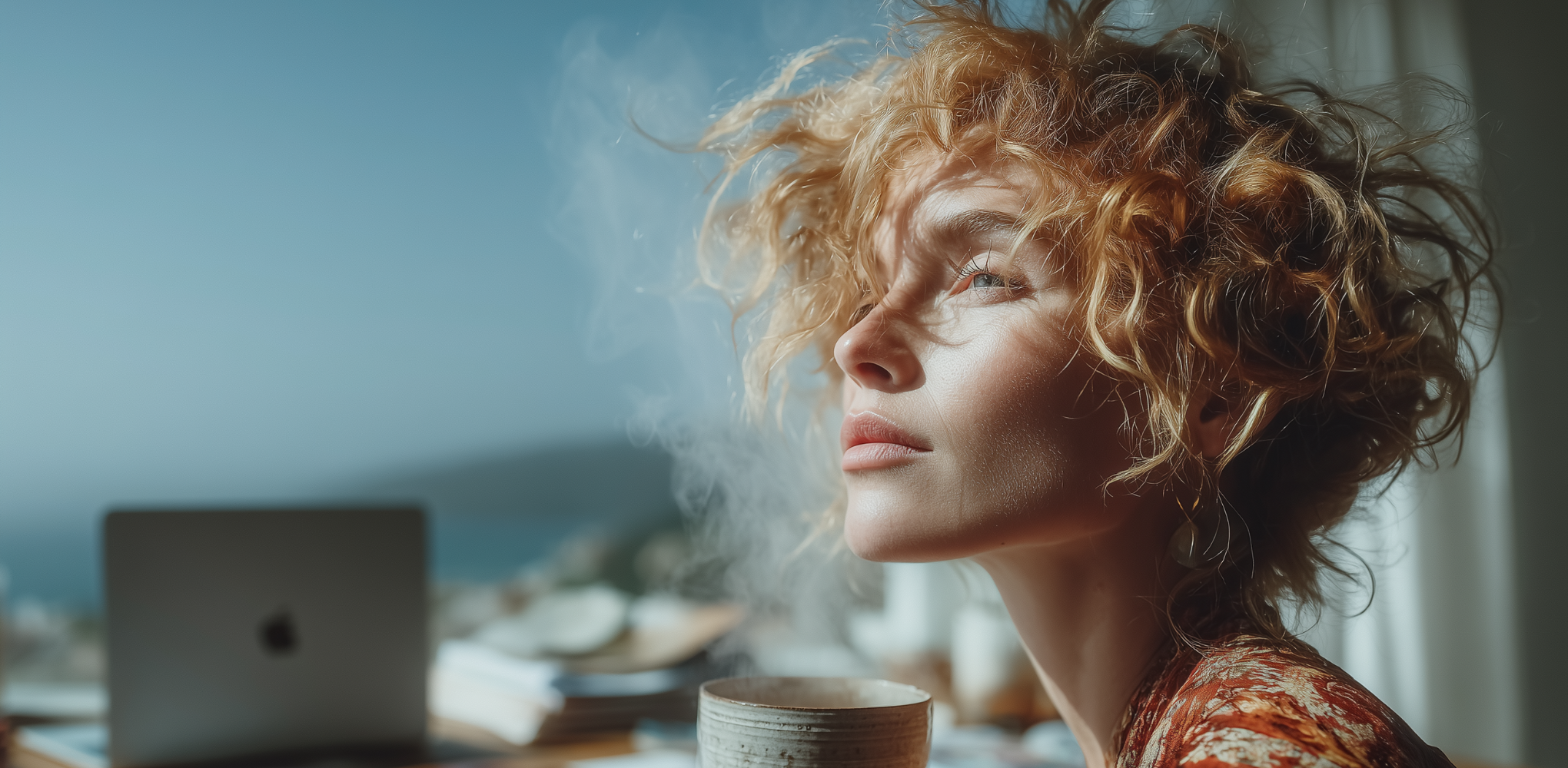 Woman with curly hair taking a peaceful breath by window with laptop and tea cup in home workspace