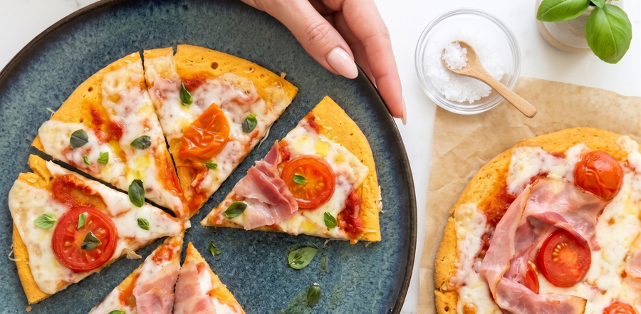 A woman's hand taking a slice of gluten-free red lentil pizza from a blue ceramic plate. The high-protein pizza features a thin, smooth crust topped with melted cheese, tomato sauce, and fresh basil, resting on a white marble countertop.